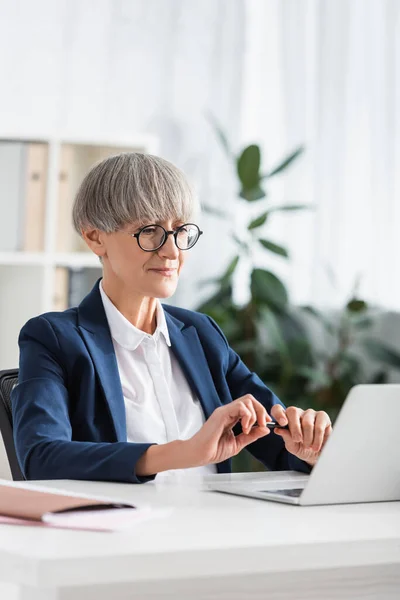 Líder de equipo de mediana edad en gafas mirando a la computadora portátil mientras sostiene la pluma - foto de stock