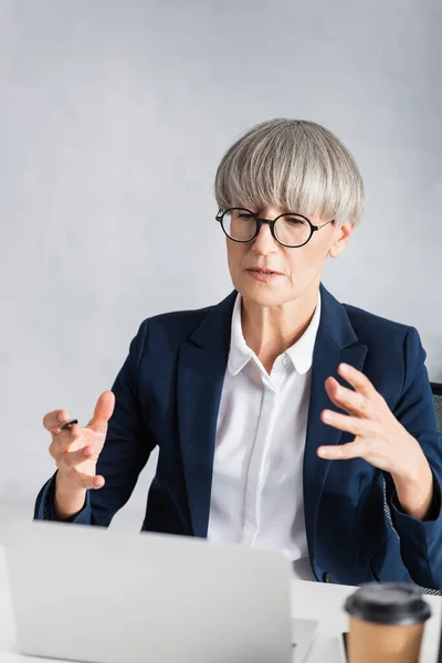 Middle aged team leader in glasses talking during video call, gesturing and holding pen — Stock Photo