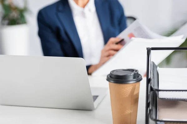 Бумажная чашка возле ноутбука на столе с бизнесвумен на размытом фоне — стоковое фото