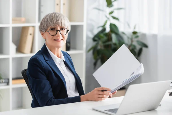 Happy team leader in glasses holding folder with documents near laptop on desk — Stock Photo