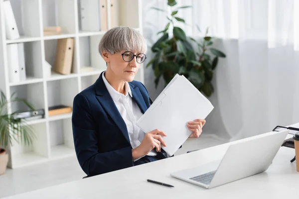 Mature team leader in glasses holding folder and looking at laptop on desk — Stock Photo