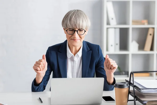 Glücklich gereifter Teamleiter mit Brille, der Daumen nach oben zeigt, während er auf Laptop schaut — Stockfoto