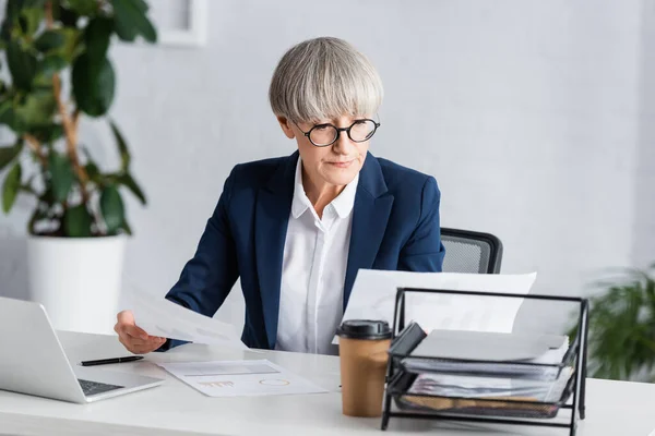 Chef d'équipe d'âge moyen dans des lunettes tenant des papiers avec des graphiques et des graphiques près d'un ordinateur portable sur le bureau — Photo de stock