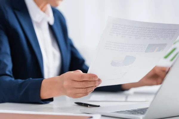 Cropped view of middle aged team leader holding papers with charts and graphs near laptop on desk — Stock Photo