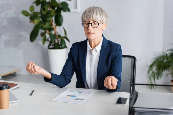 Middle aged team leader in glasses meditating near paper with charts and graphs on desk — Stock Photo