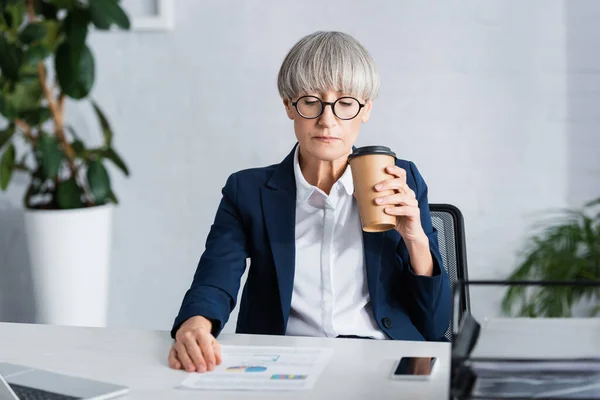 Chef d'équipe d'âge moyen dans des lunettes tenant tasse en papier près des papiers avec des graphiques et des graphiques sur le bureau — Photo de stock