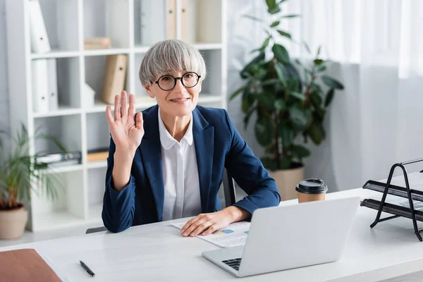 Happy middle aged team leader in glasses waving hand while looking at camera in office — Stock Photo