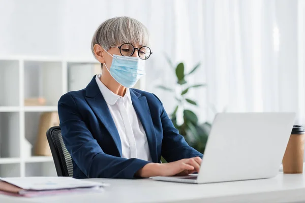 Leader del team di mezza età in occhiali e maschera medica utilizzando il computer portatile in ufficio — Foto stock