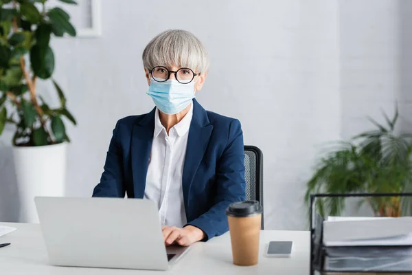 Líder de equipe de meia-idade em óculos e máscara médica usando laptop perto de smartphone e copo de papel na mesa — Fotografia de Stock