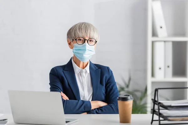 Middle aged team leader in glasses and medical mask sitting with crossed arms near laptop and paper cup on desk — Stock Photo