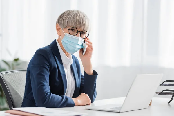 Middle aged team leader in glasses and medical mask talking on mobile phone near laptop on desk — Stock Photo