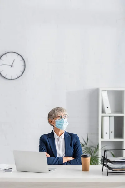 Зрелый руководитель группы в очках и медицинской маске сидит со скрещенными руками рядом с ноутбуком и бумажной чашкой на столе — стоковое фото