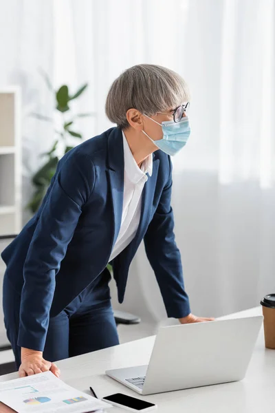 Maturo team leader in occhiali e maschera medica in piedi vicino al tavolo — Foto stock