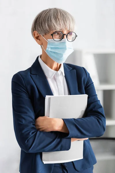 Mature chef d'équipe en lunettes et masque médical tenant dossier dans le bureau — Photo de stock
