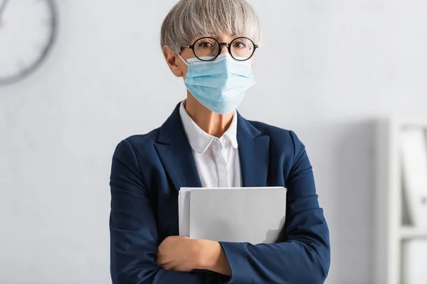 Líder de equipe madura em óculos e máscara médica segurando pasta enquanto olha para a câmera — Fotografia de Stock