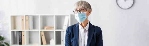 Líder de equipe madura em óculos e máscara médica olhando para longe no escritório, banner — Fotografia de Stock