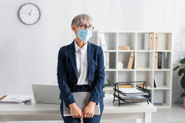 Líder del equipo maduro en gafas y carpeta de retención de máscara médica mientras está cerca del lugar de trabajo - foto de stock