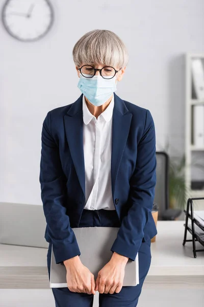 Chef d'équipe d'âge moyen en lunettes et masque médical tenant dossier tout en se tenant près du lieu de travail — Photo de stock