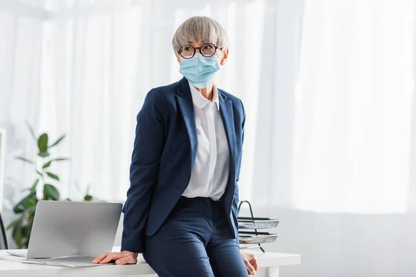 Líder de equipo maduro en gafas y máscara médica apoyada en el escritorio con computadora portátil en la oficina - foto de stock