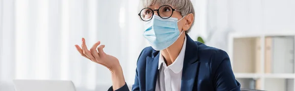 Зрелый лидер команды в очках и медицинской маске жестом, глядя вверх в офисе, баннер — стоковое фото