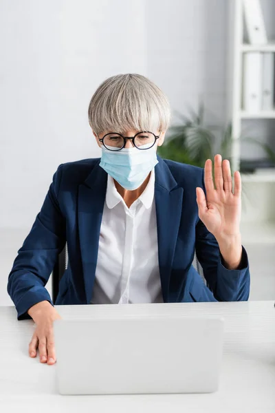 Mature team leader in glasses and medical mask waving hand while having video call in office — Stock Photo