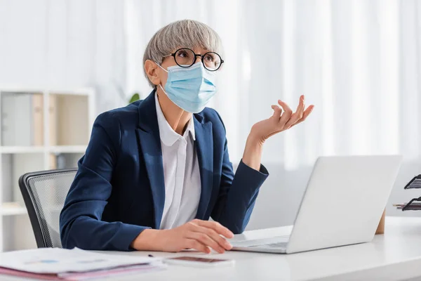 Líder de equipe madura em óculos e máscara médica gestos enquanto olha para cima no escritório — Fotografia de Stock