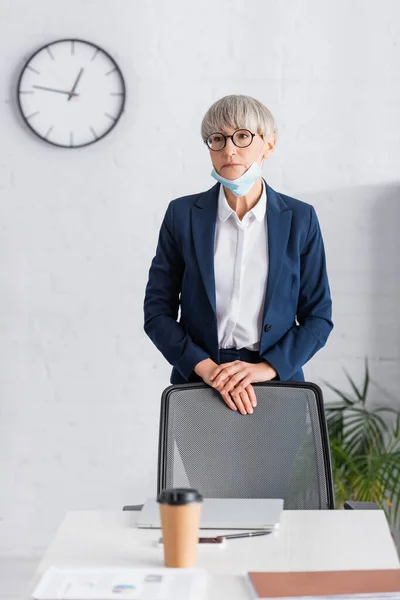 Mature chef d'équipe en lunettes et masque médical debout près du bureau avec tasse en papier au premier plan flou — Photo de stock