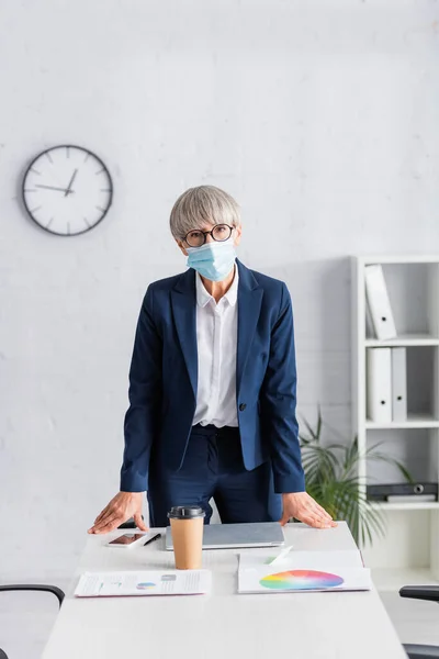 Лідер команди середнього віку в окулярах та медичній масці, що стоїть біля робочого місця в сучасному офісі — стокове фото