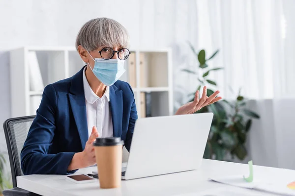 Mature team leader in glasses and medical mask gesturing near laptop on desk — Stock Photo