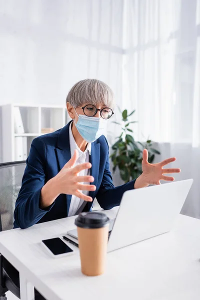 Mature chef d'équipe en lunettes et masque médical geste lors de l'appel vidéo au bureau — Photo de stock