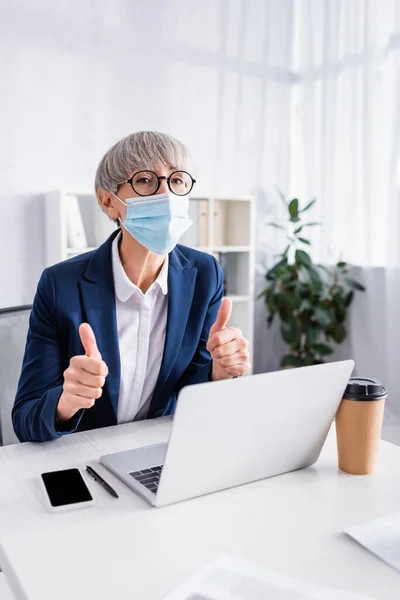 Mature team leader in glasses and medical mask showing thumbs up near gadgets on desk — Stock Photo