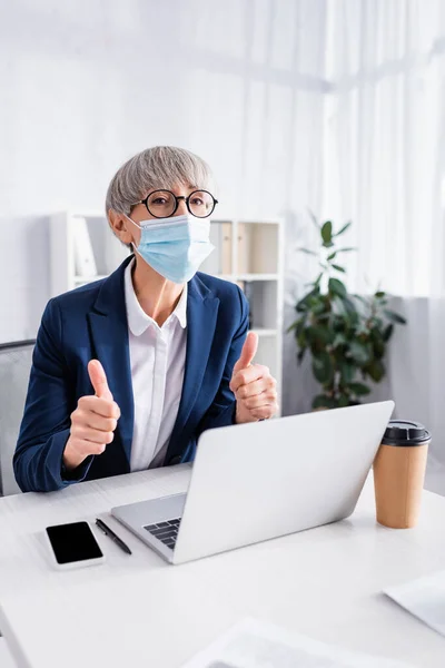 Reifer Teamleiter mit Brille und medizinischer Maske zeigt Daumen hoch in der Nähe von Gadgets auf dem Schreibtisch — Stockfoto