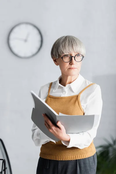 Mature team leader in glasses holding folder with documents and looking away — Stock Photo