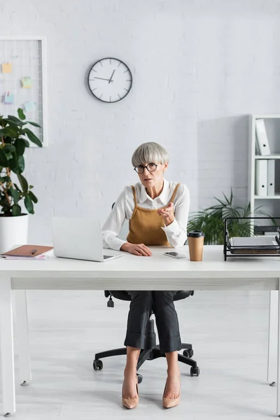 Líder de equipo de mediana edad en gafas sentadas en el escritorio y apuntando con la mano - foto de stock