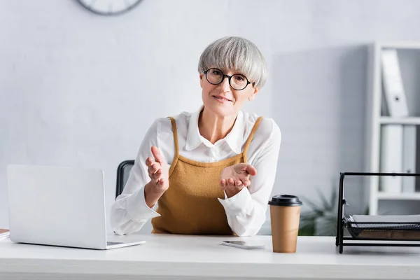 Middle aged team leader in glasses gesturing while looking at camera near gadgets on desk — Stock Photo