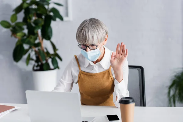 Líder de equipo de mediana edad en gafas y máscara médica saludando de la mano mientras tiene video chat en la oficina - foto de stock