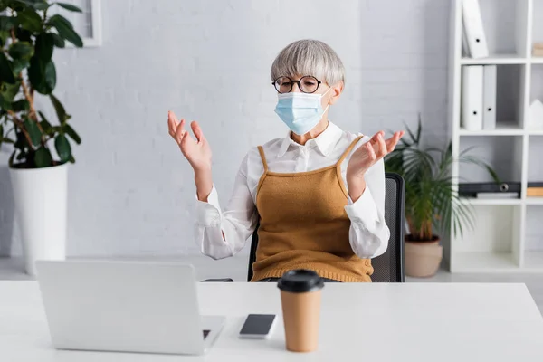 Middle aged team leader in glasses and medical mask gesturing during video call — Stock Photo