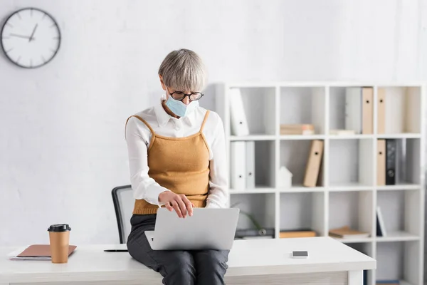 Chef d'équipe d'âge moyen dans les lunettes et le masque médical à l'aide d'un ordinateur portable assis sur le bureau — Photo de stock