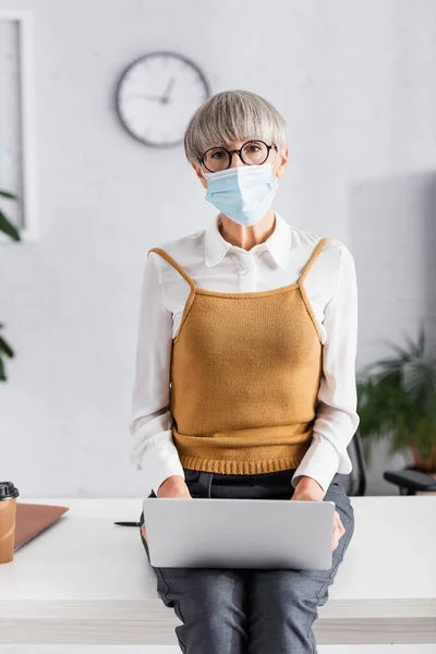 Лідер команди середнього віку в окулярах і медичній масці, використовуючи ноутбук, сидячи на столі в офісі — стокове фото