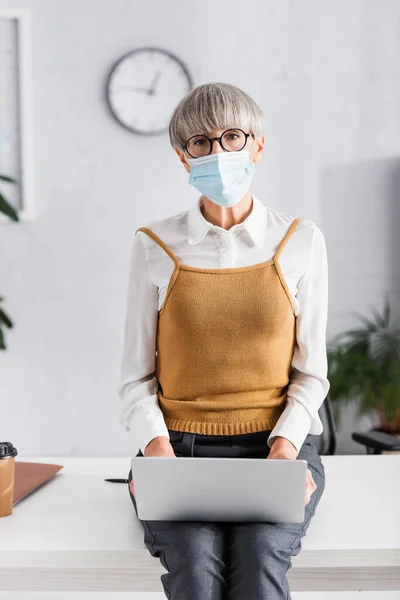 Leader del team di mezza età in occhiali e maschera medica utilizzando il computer portatile mentre seduto sulla scrivania in ufficio — Foto stock