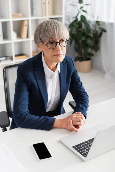 Líder de equipo de mediana edad en gafas sentadas con las manos cerradas cerca de los dispositivos en el escritorio - foto de stock