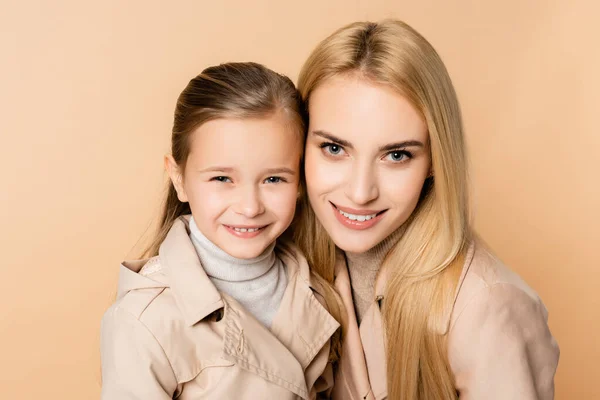 Allegra madre bionda e figlia felice in cappotti guardando la fotocamera isolata sul beige — Foto stock