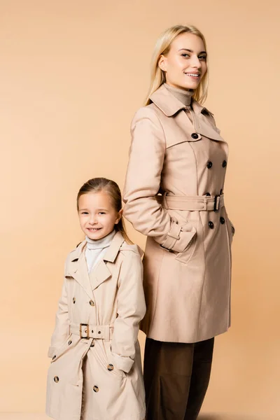 Stilvolle Mutter und Tochter in Trenchcoats lächelnd, während sie mit Händen in Taschen isoliert auf beige stehen — Stockfoto