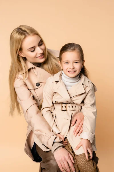 Happy mother looking at smiling daughter in trench coat isolated on beige — Stock Photo