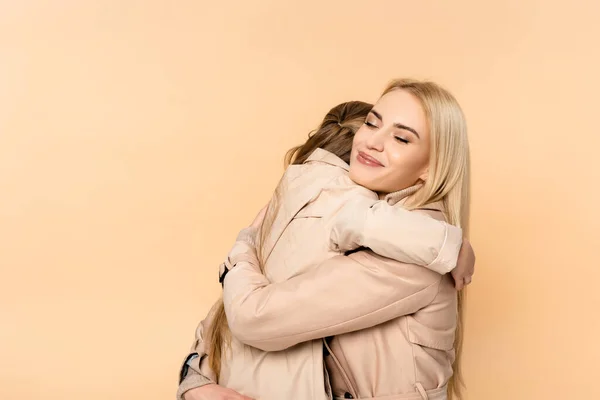 Happy blonde mother hugging daughter in trench coat isolated on beige — Stock Photo