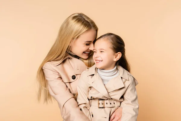 Gioiosa madre bionda che abbraccia felice figlia in trench isolato su beige — Foto stock