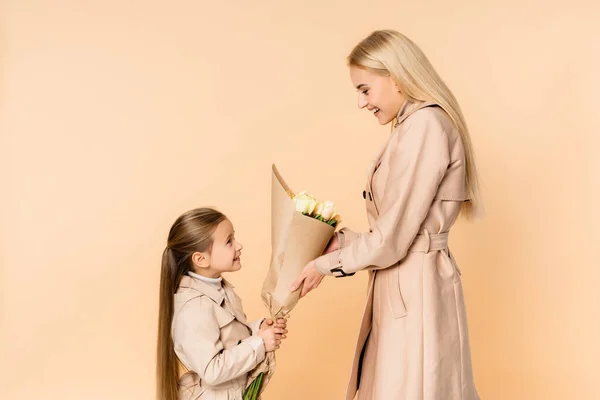 Vista laterale del bambino allegro che dà fiori alla madre felice l '8 marzo isolato sul beige — Foto stock