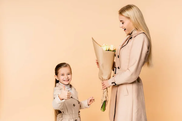 Criança alegre mostrando polegar para cima perto mãe espantada segurando flores em 8 marcha isolado no bege — Fotografia de Stock