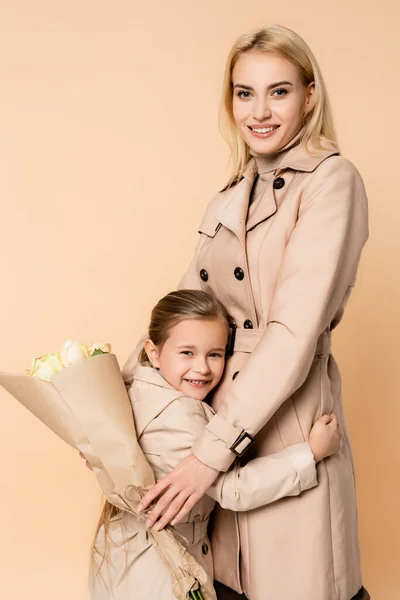 Freudige Mutter hält Blumen in der Hand und umarmt fröhliche Tochter isoliert auf beige — Stockfoto