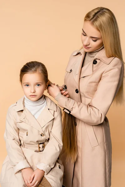 Rubia madre tejiendo trenza en hija aislado en beige - foto de stock